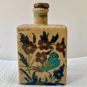 May include: A rectangular ceramic bottle with a cork stopper. The bottle is beige with a floral and bird design in shades of blue, burgundy, and green. The bottle has a cracked, aged appearance.