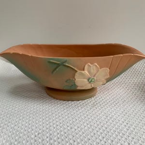May include: A peach-colored ceramic bowl with a floral design. The bowl has a wide, fluted rim and a round base. A cream-colored flower with green accents is on the side of the bowl. The bowl is likely vintage and suitable for decorative use.