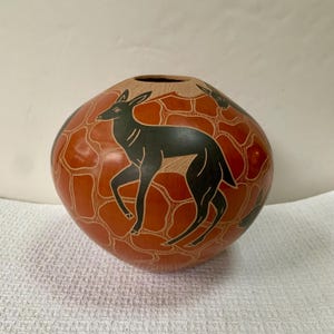 May include: A round, terracotta-colored ceramic vase featuring a hand-etched pattern of dark black deer silhouettes. The surface is covered in a grid of incised lines that create a textured, mosaic-like effect across the entire rounded body.