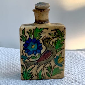 May include: A rectangular ceramic bottle with a stopper, hand-painted with a bird, blue flowers, and green foliage. The bottle has a beige background and is likely a decorative item.