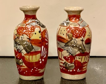 May include: Two small, hand-painted ceramic vases. The vases have a beige base, red, yellow, and gray designs, and a beige rim. The design includes abstract shapes and patterns. The vases are approximately 4 inches tall.