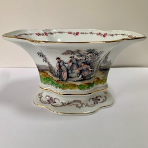 May include: A white porcelain bowl with a scalloped rim and gold trim. The exterior features a colorful hand-painted scene of three figures in a landscape with trees, while the base is decorated with a delicate floral vine pattern in purple and green.