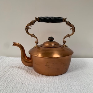 May include: A copper tea kettle with a rounded body and a curved spout. The kettle features an ornate brass handle with intricate scrollwork and a black ribbed grip. A matching lid with a small black knob sits on top of the main copper vessel.