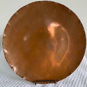 May include: A large, round copper plate with a hammered finish. The plate has a slightly scalloped edge and a rich, warm brown color. It is displayed on a black metal stand, showcasing its reflective surface.