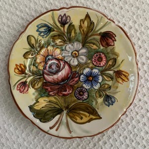 May include: A decorative ceramic plate with a floral design. The plate features a bouquet of pink, blue, yellow, and white flowers with green leaves. The plate has a scalloped edge and a brown rim.