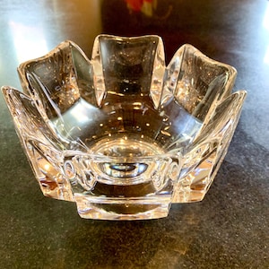 May include: A clear, decorative glass bowl with a unique, flower-like design. The bowl has a series of pointed edges that create a starburst effect. The bowl is sitting on a dark surface, reflecting light.