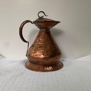 May include: A copper pitcher with a hammered texture, featuring a curved handle and a lid with a decorative swirl. The pitcher has a wide base and a bulbous body, showcasing a vintage aesthetic.