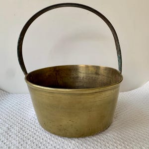 May include: A vintage brass bucket with a curved handle. The bucket has a rounded base and a slightly flared rim. The handle is dark and arched. The brass has a warm, golden hue, showing signs of age and use.
