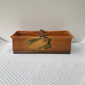 May include: A rectangular ceramic planter in shades of brown and tan, featuring a decorative pine cone and needle design. The planter has a handle made to look like a branch. The planter is 12 inches long.