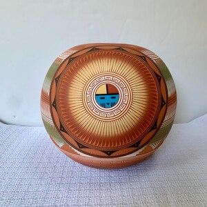 May include: A round ceramic vessel featuring a central circular motif with a stylized face in blue, black, and tan. The surface is decorated with intricate radiating lines, geometric patterns, and bands of green, brown, and cream colors.
