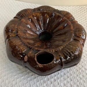 May include: A brown ceramic mold with a fluted design and a central hole. The mold has a glossy finish and a decorative, scalloped edge. It features a smaller hole on the side. The object is likely used for baking or food preparation.
