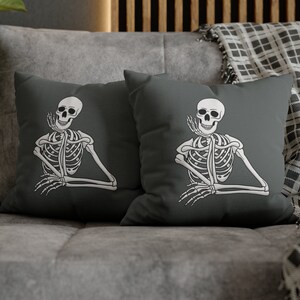 May include: Two square decorative pillows with a dark gray background. Each pillow features a white skeleton illustration in a relaxed pose. The skeleton is smiling and has its hand up near its face. The pillows are on a gray couch.