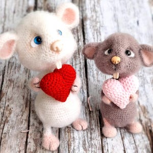 May include: Two crocheted mouse toys holding heart-shaped crocheted gifts. The mouse on the left is white with blue eyes and is holding a red heart. The mouse on the right is gray with brown eyes and is holding a pink heart.