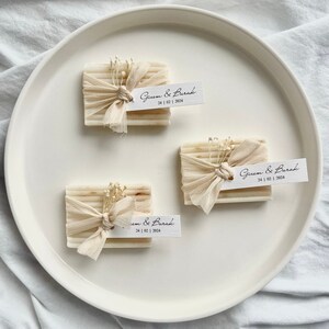 May include: Three small white bars of soap wrapped in beige fabric with white flowers and a small white tag with the text "Gizem & Burak 24 | 02 | 2024" on each.
