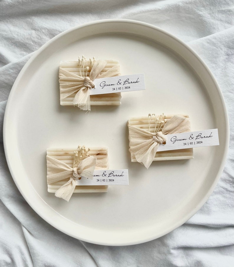 May include: Three small white bars of soap wrapped in beige fabric with white flowers and a small white tag with the text "Gizem & Burak 24 | 02 | 2024" on each.