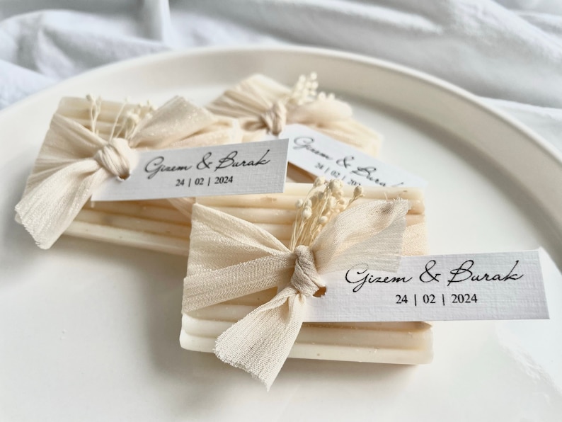 May include: Three small white soap bars wrapped in white fabric with a white flower and a tag that says "Gizem & Burak 24|02|2024".