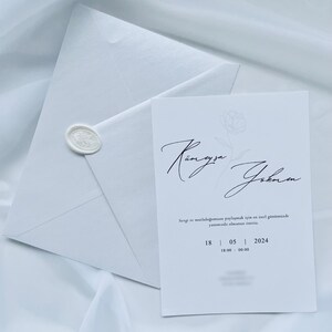 Puede incluir: Un conjunto de invitaci&oacute;n de boda blanco con tarjeta y sobre. La tarjeta tiene una escritura elegante y detalles florales, con la fecha 18/05/2024. El sobre est&aacute; sellado con un sello de cera, todo sobre una tela blanca suave.