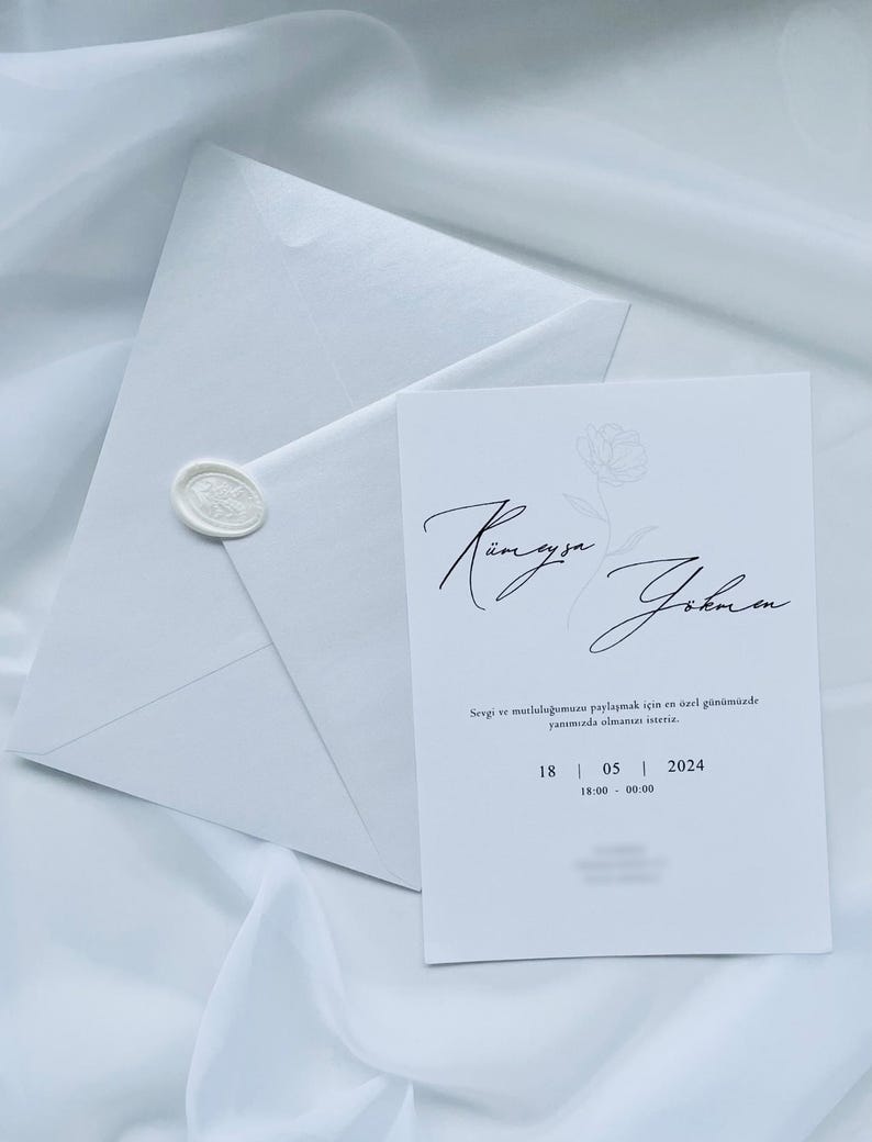 Puede incluir: Un conjunto de invitaci&oacute;n de boda blanco con tarjeta y sobre. La tarjeta tiene una escritura elegante y detalles florales, con la fecha 18/05/2024. El sobre est&aacute; sellado con un sello de cera, todo sobre una tela blanca suave.