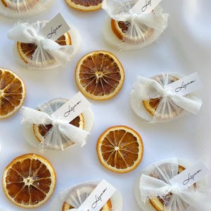 Pode incluir: Rodelas de laranja secas envoltas em tecido branco com uma pequena etiqueta branca que diz "Liam" em cada uma. As rodelas de laranja est&atilde;o dispostas sobre uma superf&iacute;cie branca.