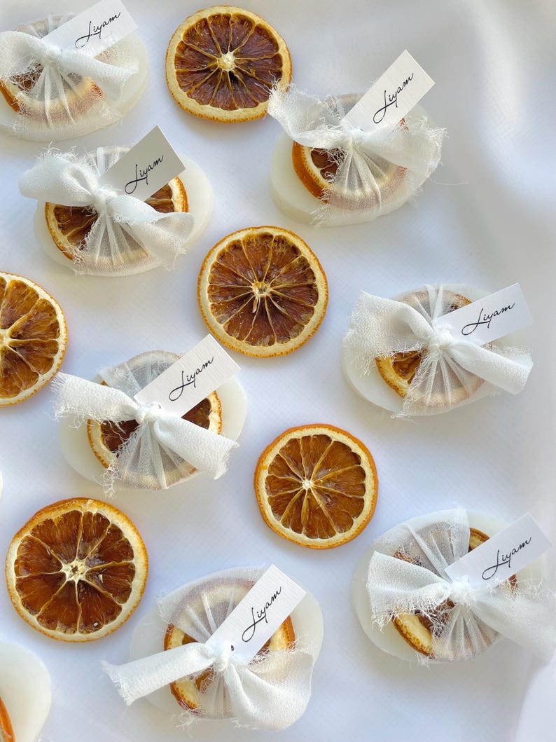 Pode incluir: Rodelas de laranja secas envoltas em tecido branco com uma pequena etiqueta branca que diz "Liam" em cada uma. As rodelas de laranja est&atilde;o dispostas sobre uma superf&iacute;cie branca.