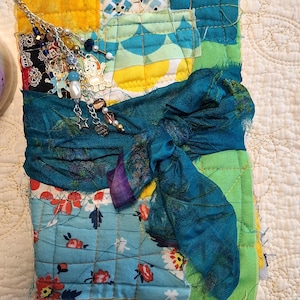 May include: A rectangular quilted fabric journal cover featuring a patchwork of yellow, teal, and floral patterned cotton squares. A dark teal silk ribbon is tied in a bow around the centre, adorned with a silver chain charm featuring beads and a small cartoon dog figure.