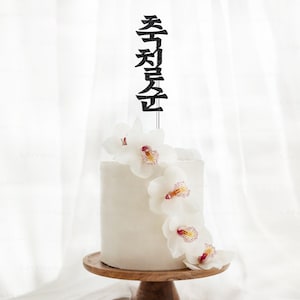May include: White cake with white orchid flowers and a black cake topper that says "Happy Birthday" in Chinese characters.