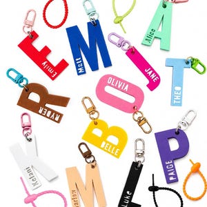 May include: A collection of colourful acrylic letter keychains in various shades including red, blue, pink, yellow, white, black, and purple. Each letter features a personalised name printed in white or black, attached to a metal clasp or plastic loop.