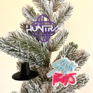 May include: Three Christmas tree ornaments hanging on a flocked artificial tree. The ornaments include a purple and white "HUNTRAX" logo, a black hat, and a "Skit Boys" logo in blue and pink.