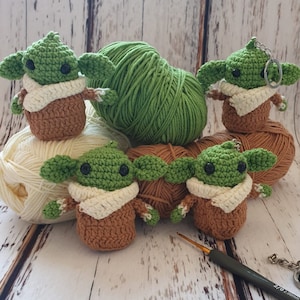 May include: Five crocheted green and brown baby Yoda figures with white yarn scarves. The figures are sitting on balls of yarn in green and white. A crochet hook and a key chain are also in the image.