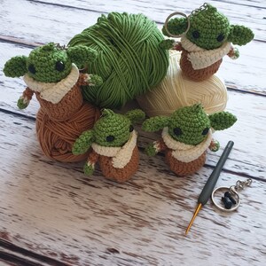 May include: Five crocheted green and brown Yoda-like figures with white accents. The figures are sitting on balls of yarn in green, white, and brown. A gray crochet hook and a silver key chain are also in the image.
