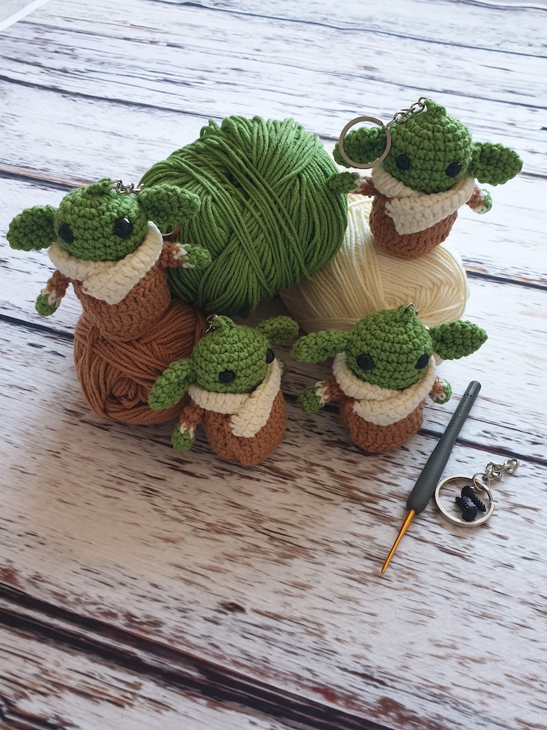 May include: Five crocheted green and brown Yoda-like figures with white accents. The figures are sitting on balls of yarn in green, white, and brown. A gray crochet hook and a silver key chain are also in the image.