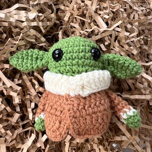 May include: A crocheted green and brown plush toy with large black eyes. The toy is wearing a white crocheted collar and a brown crocheted jacket.