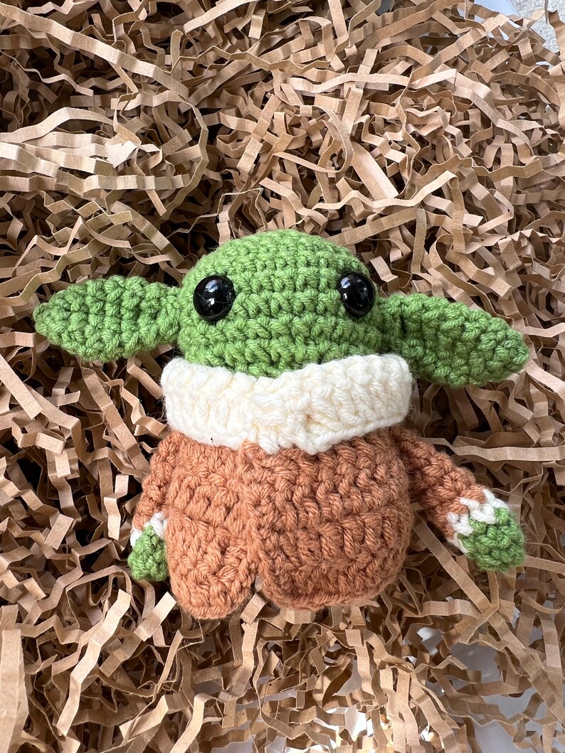 May include: A crocheted green and brown plush toy with large black eyes. The toy is wearing a white crocheted collar and a brown crocheted jacket.