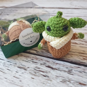 May include: A green and brown crocheted plush toy in the shape of a character from the Star Wars franchise, wearing a white scarf. The toy is in a clear plastic box with green and brown paper shreds. The box has a sticker that says "handmade with love".