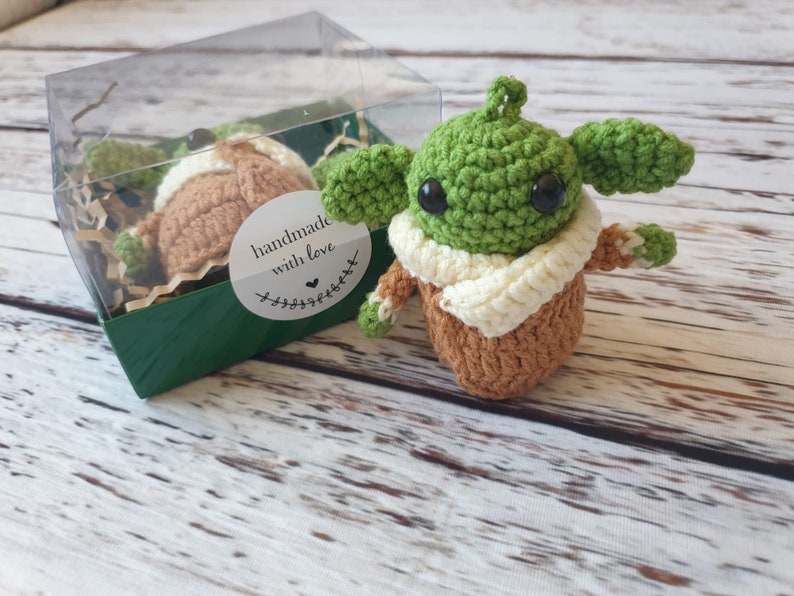 May include: A green and brown crocheted plush toy in the shape of a character from the Star Wars franchise, wearing a white scarf. The toy is in a clear plastic box with green and brown paper shreds. The box has a sticker that says "handmade with love".