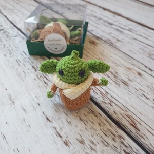 May include: A small green crocheted Yoda character with brown eyes and a tan crocheted robe. The character is sitting on a white wooden surface.