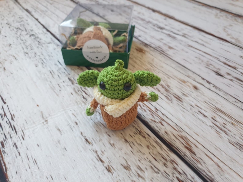 May include: A small green crocheted Yoda character with brown eyes and a tan crocheted robe. The character is sitting on a white wooden surface.