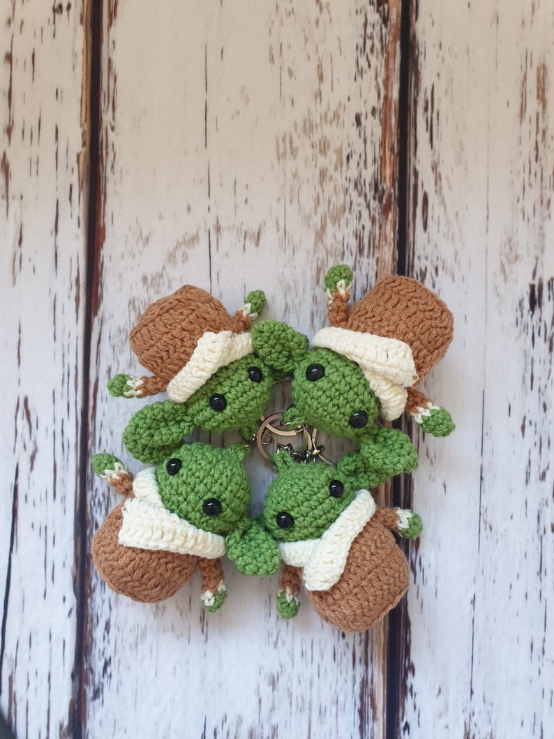 May include: Four crocheted green and brown baby Yoda keychains with white trim. The keychains are arranged in a circle on a white wooden background.
