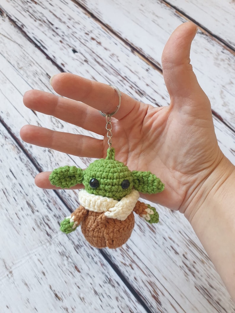 May include: Green crocheted keychain with a brown coat and a white scarf. The keychain is shaped like a popular fictional character.