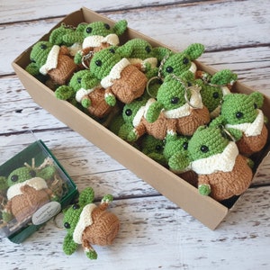 May include: A box filled with crocheted green and brown baby Yoda keychains. The keychains are arranged in a box with a brown cardboard exterior.