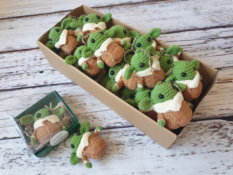 May include: A box filled with crocheted green and brown baby Yoda keychains. The keychains are arranged in a box with a brown cardboard exterior.