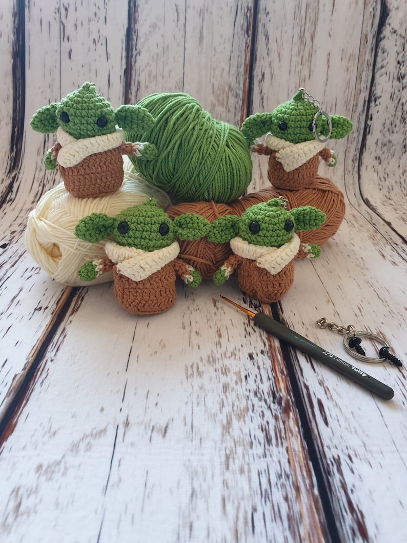 May include: Five crocheted green and brown baby Yoda figures with white yarn scarves. The figures are sitting on balls of yarn in green and white. A crochet hook and a key chain are also in the image.