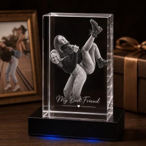 May include: A rectangular clear glass block featuring a 3D laser-etched image of two people embracing, with one person being carried on the other's back. The text "My Best Friend" is etched below the figures, resting on a black rectangular base.