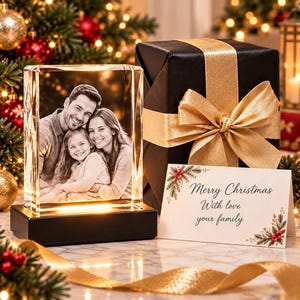 May include: A rectangular glass block featuring a laser-etched black and white photograph of a family, resting on a black light-up base. Next to it is a black gift box with a gold ribbon and a white card with the text Merry Christmas With love your family.