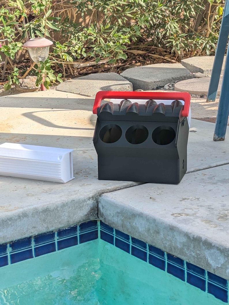 May include: Black and red plastic container with four round openings and a red handle. The container is sitting on a concrete surface next to a swimming pool.