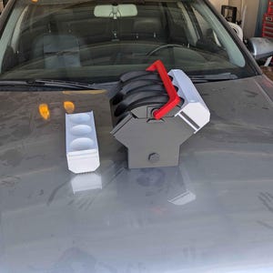 May include: A white and red 3D printed car engine model with a black base. The model is sitting on a gray car hood. There is a white 3D printed cup holder next to the engine model.