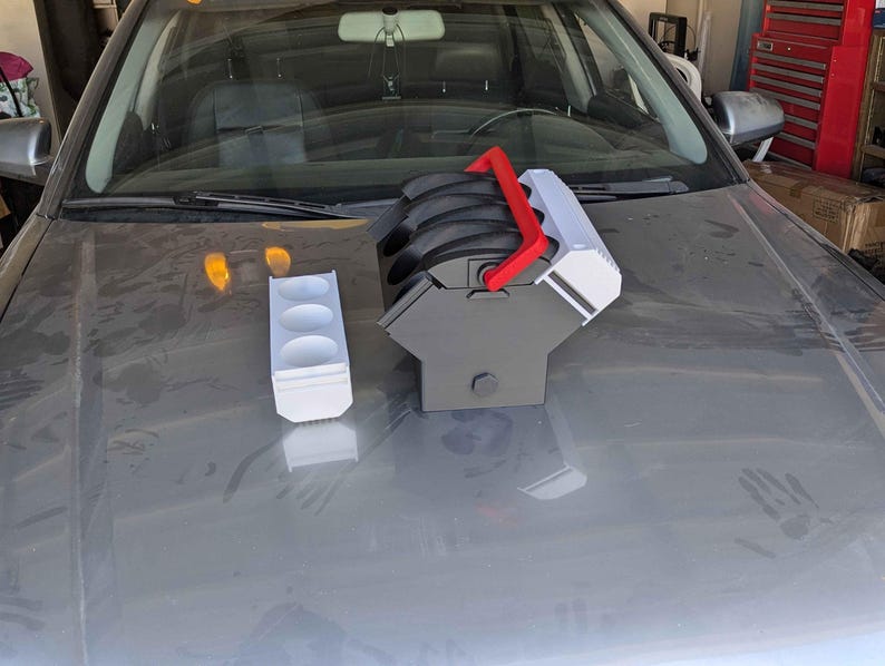 May include: A white and red 3D printed car engine model with a black base. The model is sitting on a gray car hood. There is a white 3D printed cup holder next to the engine model.