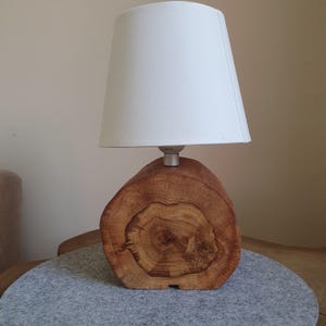 Könnte beinhalten: Eine Tischlampe mit einem Sockel aus einem natürlichen Holzstamm mit sichtbaren kreisförmigen Maserungen und einem glatten, hellen, zylindrischen Stofflampenschirm. Die Lampe hat eine silberfarbene Metallfassung, die den Holzsockel mit dem Schirm verbindet.