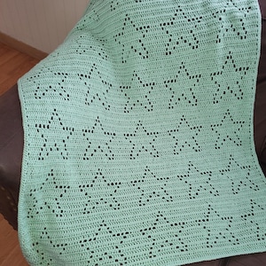 May include: A light green crochet blanket with a star pattern. The blanket is folded and draped over a brown leather chair. The text "AnnMossCrochet" is visible in the lower right corner of the image.