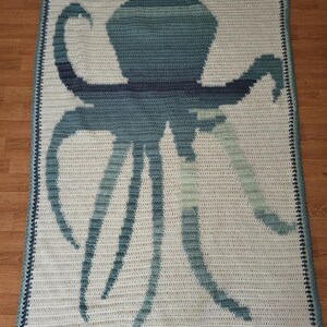 May include: A crocheted blanket featuring a large octopus design in shades of blue and green against a white background. The blanket has a dark blue border and the text "AnnMossCrochet" at the bottom.
